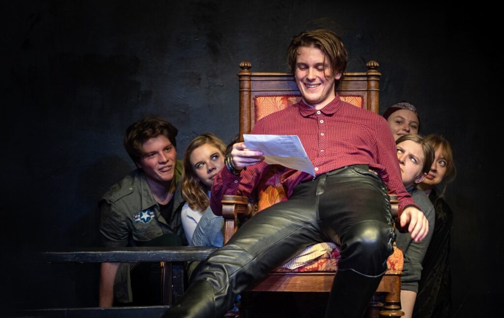 Schauspielschüler*innen auf der Bühne bei einer Szene mit starkem Ensemble-Fokus – ein Darsteller sitzt auf einem Thronstuhl und liest eine Rolle, während die anderen gespannt und ausdrucksstark reagieren. Symbolbild für Schauspielmethoden nach Stanislawski, Michael Chekhov und Stella Adler, die auf Ensemblearbeit, Imagination und Figurenanalyse setzen.